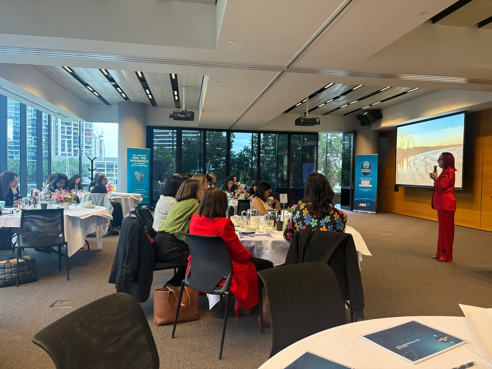 Tracy Sheen presenting in a red suit at a corporate function, audience seated at round tables