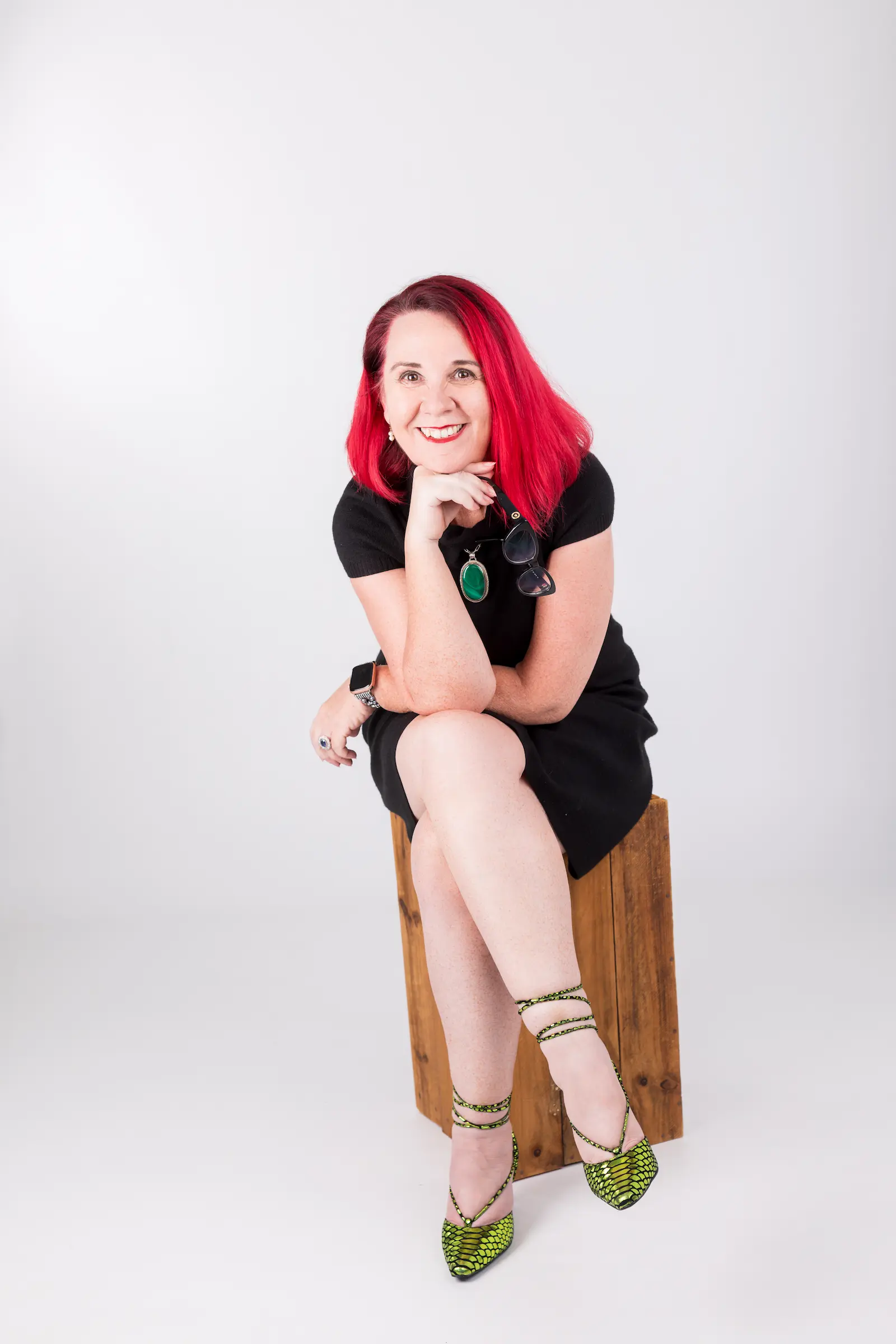 Tracy Sheen seated on a wooden box in a black dress, holding her glasses and smiling